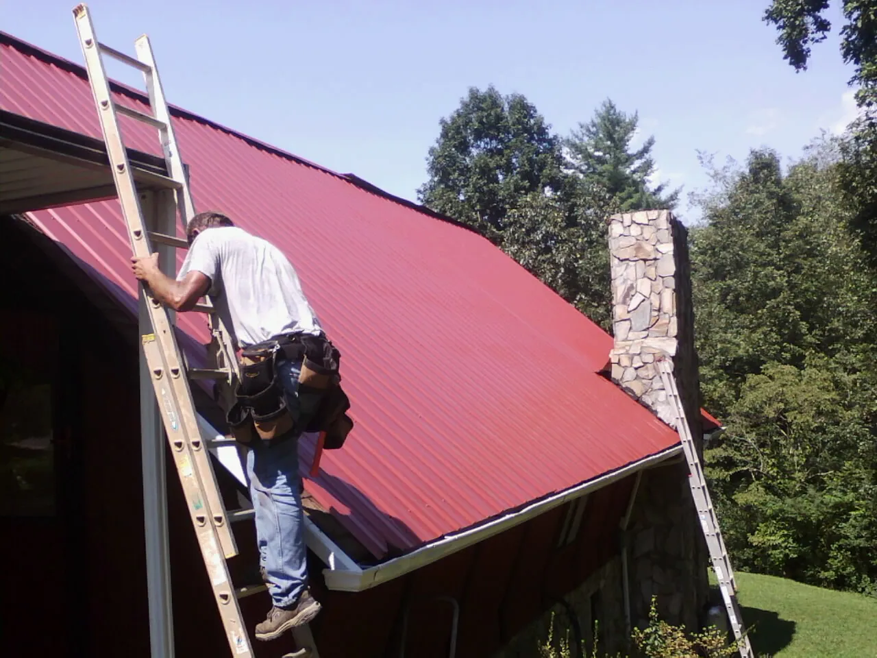 Roofer on a ladder beside a red metal roof and stone chimney — Soffit & Fascia Repair by Roofing Brothers in Foothill Ranch